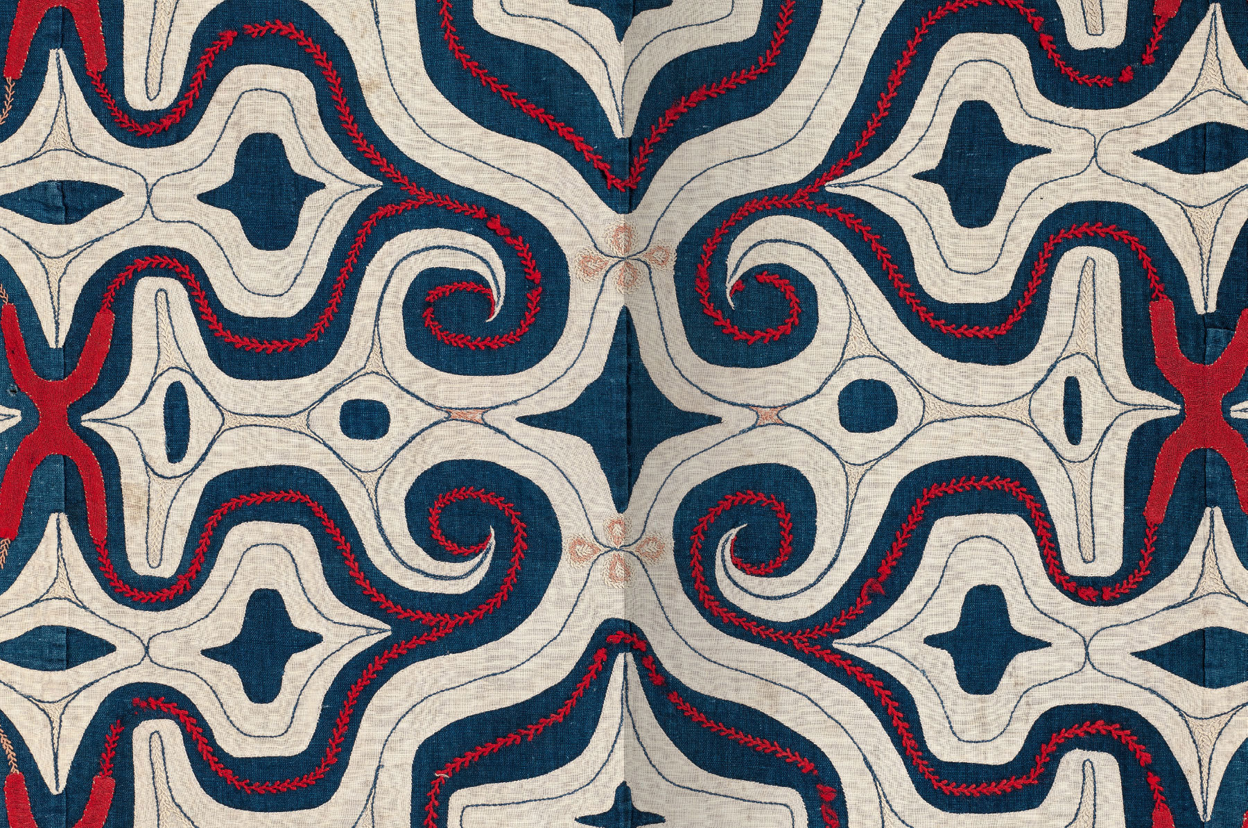 Textiles of Japan book spread showing a detail of a red, blue, and white kaparamip robe. Embroidery is used to create abstract designs as well as representational images of four-petaled flowers